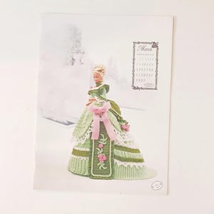 May include: A doll wearing a green and white crocheted gown with a pink bow and floral accents. The doll is on a calendar page with the month of March and the year 1997. The background is a blurred winter scene.