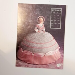 May include: A calendar page featuring a doll dressed in a crocheted gown. The doll's dress is primarily gray with pink accents and a pink skirt. The calendar shows the month of September.