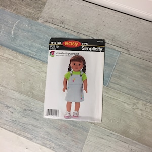 May include: A Simplicity sewing pattern for a doll's dress. The pattern cover features a doll wearing a light blue overall dress with colorful embroidered details and a green shirt. The text on the cover reads "It's So Easy" and "create-it-yourself."