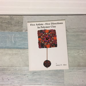 May include: A book titled "Five Artists - Five Directions In Polymer Clay" features a colorful, square clock design on the cover. The clock face is a vibrant mix of red, orange, blue, and black patterns. The book is by Jamey D. Allen.