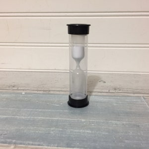 May include: A clear hourglass with black caps at the top and bottom. The hourglass contains white sand. The hourglass is standing on a light blue and gray wooden surface.