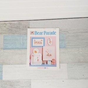 May include: A book cover titled "Bear Parade" featuring cross-stitch designs of teddy bears with balloons and gifts. The cover includes the Dimensions logo and a price tag. The color palette includes pink, blue, and white.