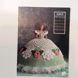 May include: A doll wearing a green and white crocheted dress with pink rose accents. The doll is on a calendar page for March. The calendar has the days of the month listed. The doll has brown hair and is wearing a green top.
