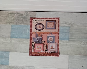 May include: A framed print featuring the text "SUNBONNET BABIES" and various cross-stitch designs. The designs include framed pictures, a doll, and a bonnet. The print has a red border with white polka dots and is displayed on a wall with a wood-like pattern.