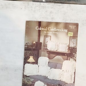 May include: A book titled "Colonial Candlewicking" by Janice Shirley. The cover features a vintage interior scene with white ruffled pillows and a sofa. The book has a Walmart price sticker.
