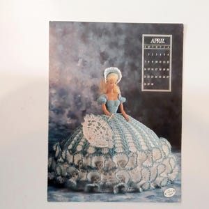 May include: A doll wearing a light blue and white crocheted gown and bonnet, holding a matching purse. The image is a calendar page for April, with a cloudy blue background. The calendar grid is in the upper right corner.