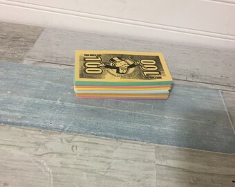 May include: A stack of Monopoly game money, featuring a yellow top bill with the Monopoly logo and the text "100 Million". The stack includes bills in various colors, such as pink, green, and blue. The game money is stacked on a wooden surface.