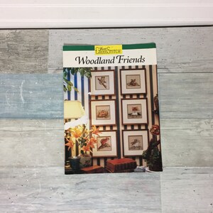 May include: A Just Cross Stitch pattern book titled "Woodland Friends" featuring framed cross-stitch samplers of woodland animals. The cover shows a display of framed samplers, a lamp, and flowers. The background is a blue and white striped wall.