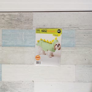 May include: A dog wearing a green dinosaur costume with yellow spikes, from a Simplicity sewing pattern. The pattern package is on a light blue and white wood-paneled surface. The text on the package reads "Sew Simple".