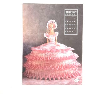 May include: A calendar page featuring a doll in a pink dress and bonnet. The dress has a layered design with intricate detailing. The calendar displays the month of February with a list of dates.