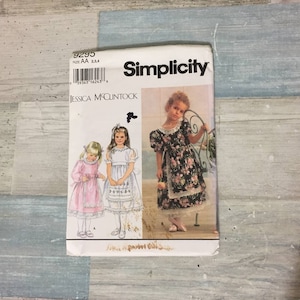 May include: A Simplicity sewing pattern featuring designs for girls' dresses. The pattern cover displays illustrations of three different dress styles, with a floral print dress and a pink dress with lace trim. The pattern is size AA 2,3,4.