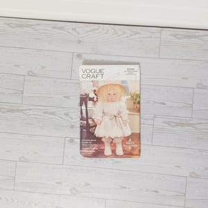 May include: A Vogue Craft pattern featuring a doll in a white dress, hat, and bloomers. The doll is standing in front of a chair and a table with flowers. The pattern is labeled "Vogue Craft" and has the number "8240".