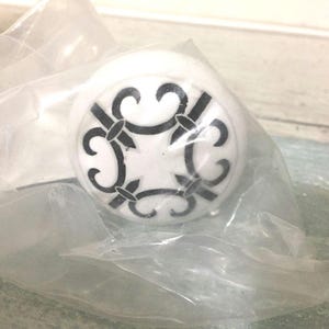 May include: A white cabinet knob with a black fleur-de-lis design. The knob is round and appears to be made of ceramic or porcelain. The design is symmetrical and centered on the knob's face. The knob is in a clear plastic bag.