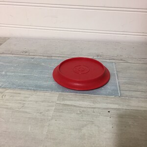 May include: A bright red, circular plastic lid with a slightly raised edge. The lid has a recessed center with a circular design. The lid is sitting on a light blue and gray wooden surface.