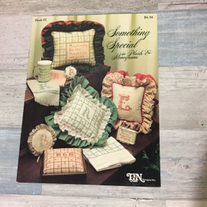 May include: A book cover titled "Something Special in Plaids & Monograms" featuring various embroidered items. These include pillows, a mug, and a small box, all with embroidered letters and designs. The color palette includes red, green, and cream.