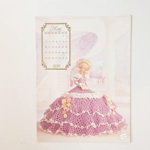 May include: A 1992 calendar page featuring a doll in a crocheted lavender dress with white trim and yellow floral accents. The calendar displays the month of May.