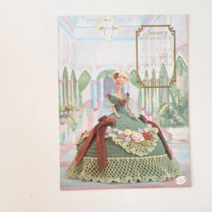 May include: A calendar page featuring a doll in a green crocheted gown with red ribbon accents and floral embellishments. The calendar is dated January 2002, set against a backdrop of an ornate interior with columns and plants.