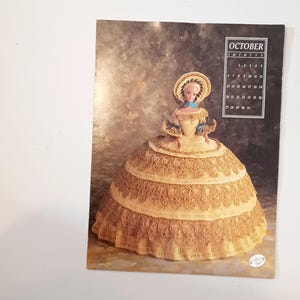 May include: A calendar page featuring a doll in a large, tiered, crocheted dress. The doll has a matching hat and a blue scarf. The calendar shows the month of October. The background is a mottled brown.