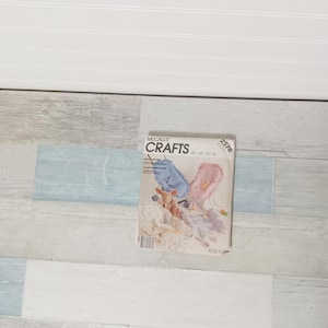 May include: A McCall's Crafts pattern booklet featuring a baby-themed sewing project. The cover displays images of a blue and pink baby item, a stuffed animal, and other craft elements. The booklet is set against a backdrop of light blue, gray, and white wood-look panels.