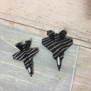 May include: Two paper airplane models of fighter jets. The jets are black with gray zebra stripes. The jets have a white nose cone. One jet is slightly smaller than the other.