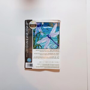 May include: A Dimensions brand cross-stitch kit featuring a dragonfly design. The kit includes a canvas with a pre-printed image of a dragonfly with white wings and a blue body, surrounded by green foliage. The kit measures 15 cm x 15 cm.
