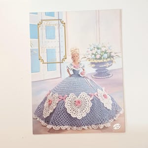 May include: A vintage Barbie doll calendar page featuring a doll in a blue crocheted gown with white lace heart accents and pink bows. The calendar displays the month of February, 1992, with a floral arrangement in the background.