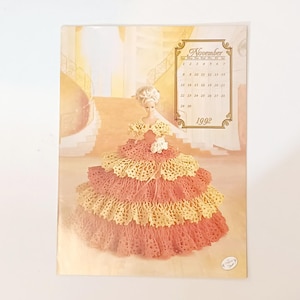 May include: A vintage calendar page featuring a doll in a layered, peach and yellow crocheted gown. The calendar is dated November 1992. The background shows a staircase and architectural details, with a soft, warm color palette.