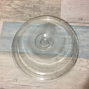 May include: A clear glass lid with a circular design. The lid has a central knob and concentric rings with a ribbed pattern. The glass is transparent, allowing a view of the surface beneath. The lid is likely for a pot or pan.