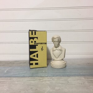May include: A cream-colored bust of Franz Schubert with the name "SCHUBERT" on the base. Beside it is a yellow and black box with the word "HALBE" and "STATUETTE" printed on it.