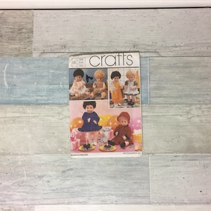 May include: A vintage crafts pattern book titled "crafts" featuring illustrations of dolls in various outfits. The cover shows dolls in dresses and outfits, with text indicating the pattern is for large dolls. The background is a light blue and white wood-like surface.