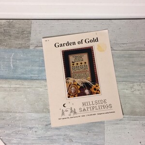 May include: A cream-colored paper with the title "Garden of Gold" and a cross-stitch design. The design includes sunflowers, text, and the alphabet. The paper is from Hillside Samplings, East Aurora, NY.