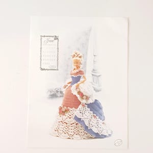 May include: A printed image featuring a doll in a vintage gown with a matching hat. The doll's dress is a combination of pink, blue, and white, with intricate lace detailing. The image also includes a calendar for the month of June.