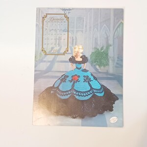 May include: A 1992 calendar page featuring a doll in a blue and black dress. The doll has blonde hair and is standing in front of a painted background. The calendar shows the month of December.