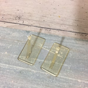 May include: Two clear, rectangular adhesive hooks with a small peg extending from the center. The hooks are made of a transparent plastic material. The background is a weathered, light blue and gray surface.