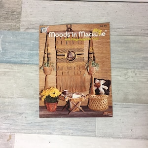 May include: A vintage magazine cover titled "Moods in Macrame" with a macrame wall hanging, hanging planters, and a woven stool. The cover features a warm color palette with brown, yellow, and green tones. The magazine showcases macrame craft projects.