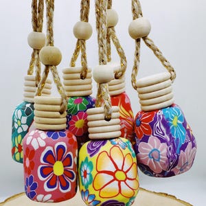 May include: A collection of colorful, floral-patterned car air fresheners. Each bottle-shaped freshener features a unique design with bright flowers in shades of pink, red, yellow, blue, and green. They hang from braided cords with wooden beads and caps.
