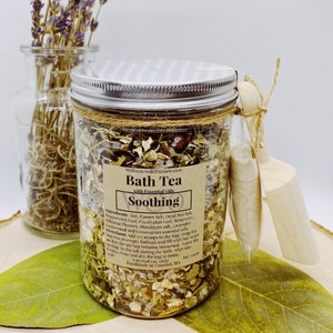 May include: A glass jar filled with a blend of dried herbs, flowers, and salts labelled "Bath Tea with Essential Oils Soothing". The label lists ingredients including oat, Epsom salt, Dead Sea salt, peppermint leaf, eucalyptus leaf, rosemary, hibiscus flower, Himalayan salt, lavender, cedarwood, and lemongrass plus essential oils. Directions for use are included on the label.