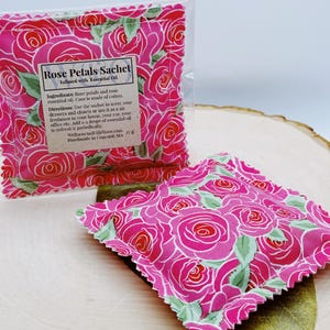 Op de afbeelding: Een roze stoffen zakje met een rozenpatroon. Het zakje is gevuld met rozenblaadjes en etherische olie. De tekst op het zakje luidt "Rose Petals Sachet Infused with Essential Oil".