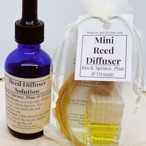 Op de afbeelding: Een klein, doorzichtig glazen flesje met een blauwe dop gevuld met een gele vloeistof. Het flesje is gelabeld "Reed Diffuser Solution Black Spruce, Pine & Orange". Het flesje zit in een klein wit organza zakje met een label waarop staat "Mini Reed Diffuser Black Spruce, Pine & Orange".