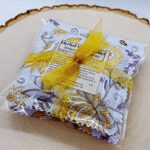 May include: A set of four lavender and chamomile infused sachets with a yellow floral pattern. The sachets are wrapped in clear plastic and tied with a yellow ribbon. The text on the label reads "Herbal Sachet Infused with Oils".