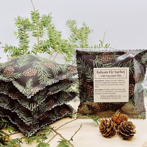 May include: A set of four balsam fir sachets with essential oils. The sachets are made of cotton fabric and feature a black and green pine cone print. The sachets are packaged in clear plastic bags. The text on the bag reads "Balsam Fir Sachet with Essential Oils".