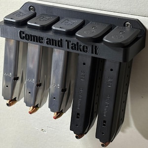 May include: A black 3D-printed magazine holder mounted on a wall, holding five magazines. The holder has the text "Come and Take It" and is designed to hold magazines. The magazines are silver and black.