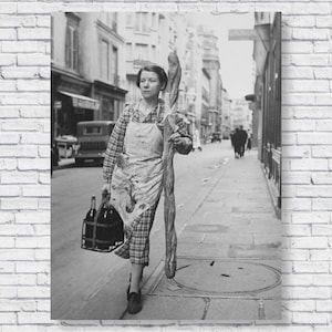 Puede incluir: Una mujer con un vestido de cuadros y un delantal lleva una caja de botellas de vino y un palo largo por una calle adoquinada de París.