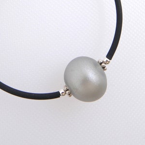 May include: A black bracelet featuring a large, oval, silver-gray bead and two small, dangling, iridescent crystal charms. The bracelet has a simple, elegant design, suitable for everyday wear or special occasions.