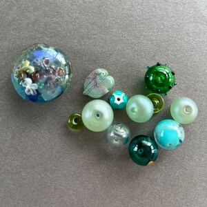 May include: A collection of green, blue, and white glass beads. The beads are various shapes and sizes, including round, heart, and star shapes. Some beads have a textured surface.
