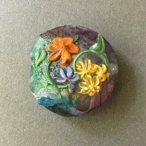 May include: A round glass bead with a floral design. The bead features orange, yellow, and blue flowers with green leaves. The background of the bead is a mix of purple, blue, and green.