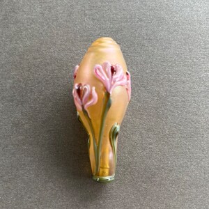 May include: A glass bead with a pale yellow and gold iridescent finish. The bead is decorated with pink and green flowers.