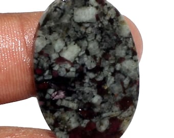 Puede incluir: Una gema ovalada con un patrón de mosaico de tonos grises, blancos y rojo oscuro. La superficie pulida revela una mezcla de texturas y colores. La gema es sostenida por un dedo.
