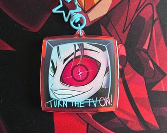 May include: A square, clear acrylic keychain with a cartoon image of a character's eye and face, with a red and white target-like iris. The keychain has a blue star-shaped clasp and the text "TURN THE TV ON!" in black.