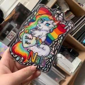 May include: A clear sticker featuring a white wolf-like character with rainbow hair and a rainbow tail. The character holds a heart with "She/Her" on it. The name "Nala" is written in rainbow colors. The sticker has a decorative border.